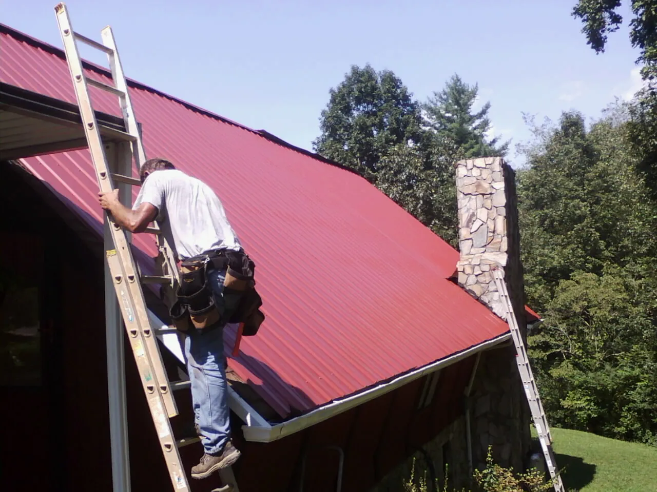 Roofer on a ladder beside a red metal roof and stone chimney — Soffit & Fascia Repair by Roofing Brothers in Kaylor